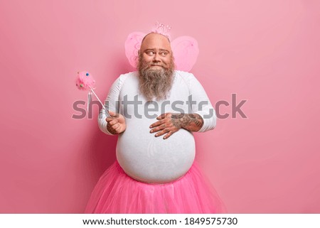 Funny bearded plump man keeps hand on fat belly holds magic wand looks happily aside entertains children at party isolated over pink background. Positive dad in carnival costume has playful mood Royalty-Free Stock Photo #1849575730