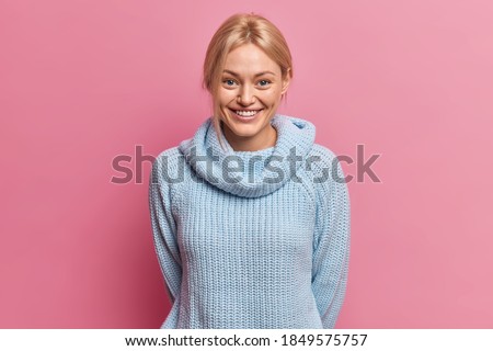 Waist up shot of pleased blonde European woman with glad tender expression keeps hands behind back dressed in knitted blue jumper expresses happy emotions isolated over pink studio background. Royalty-Free Stock Photo #1849575757