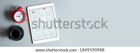 a tablet with an open calendar for 2021 year, a cup of coffee,  and a red alarm clock on a gray background. banner. space for text Royalty-Free Stock Photo #1849590988