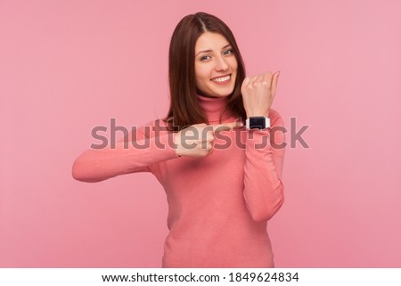 Happy positive woman with brown hair in pink sweater pointing finger on watch on her wrist looking at camera with toothy smile. Look at time! Indoor studio shot isolated on pink background Royalty-Free Stock Photo #1849624834