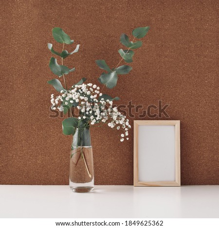 Wooden frame and vase with white flowers and eucalyptus branches, cork background. Mock up, copy space. Folk