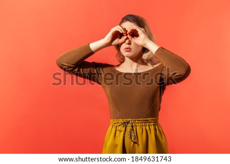 Where you? Portrait of serious blonde woman in casual brown blouse making glasses shape, looking through binoculars gesture. Indoor studio shot isolated on red background.