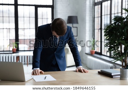 Frustrated young businessman in formal wear standing at office table with head down, feeling depressed after professional failure, company bankruptcy, money loss or unprofitable deal concluding. Royalty-Free Stock Photo #1849646155