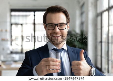 Headshot portrait of happy healthy young businessman employee in eyeglasses and formal wear holding glass of cool filtered fresh water and showing thumbs up gesture, feeling refreshed in office. Royalty-Free Stock Photo #1849647394