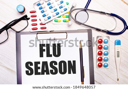 On a light wooden background there is paper with the inscription FLU SEASON, a stethoscope, colorful pills, glasses and a pen. Medical concept