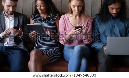 Modern technology. Group of millennial men and women of different gender and race sitting on sofa communicating by laptops smartphones pads, sharing data using wifi connection, chatting in messengers