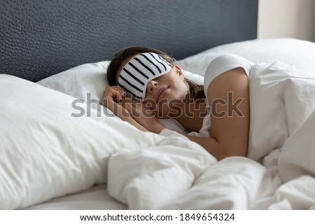 Stylish nightwear. Comfy sleeping mask helping young woman tourist traveler enjoy good healthy night sleep at new place in hotel suite room, millennial woman napping at wide king size bed in eye mask Royalty-Free Stock Photo #1849654324