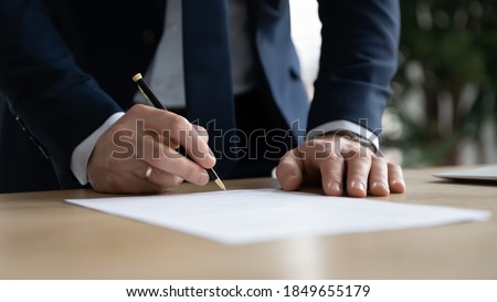 Close up young businessman standing near table with pen in hands, ready signing profitable offer agreement after checking contract terms of conditions, executive manager involved in legal paperwork. Royalty-Free Stock Photo #1849655179