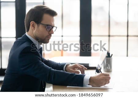 Focused young businessman in formal wear and eyeglasses working on computer, doing web research, writing notes in paper report, sitting in office, skilled ceo manager checking financial documentation.