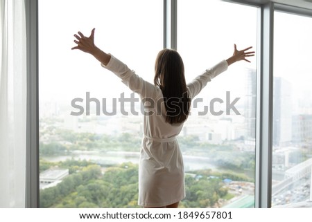 That is my world! Charming young woman in bathrobe standing at large panoramic window wall stretching hands to sides opening embraces to life enjoying splendid city panorama meeting new day, back view