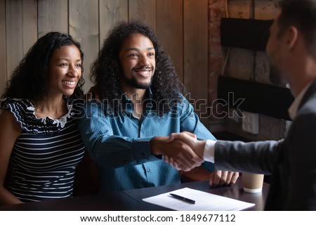 Satisfied with deal. Happy young biracial husband and wife clients and real estate broker, insurance agent, financial advisor, travel manager shake hands after making purchase and signing contract