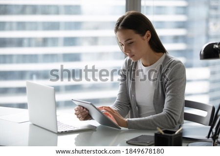 Modern office worker. Concentrated focused young businesswoman employee manager doing electronic paperwork at workplace using devices comparing checking synchronizing data at pad and laptop memory Royalty-Free Stock Photo #1849678180
