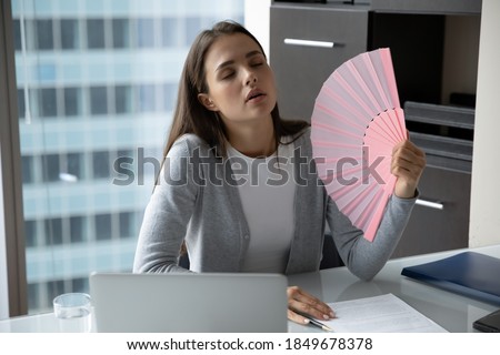What a hot day! Exhausted millennial female office worker manager employee sitting at desk waving herself with hand fan suffering from summer heat too sultry air dreaming about conditioner humidifier Royalty-Free Stock Photo #1849678378