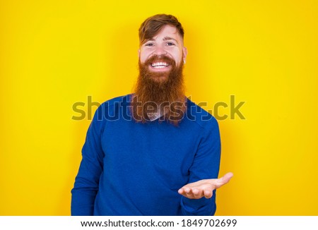 Young handsome red haired bearded Caucasian man standing against yellow wall smiling friendly offering something with open hand or handshake as greeting and welcoming. Successful business.