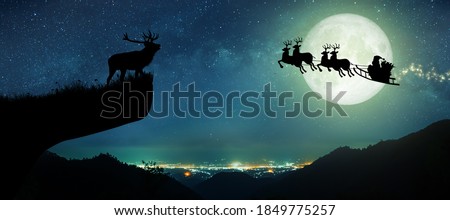 Silhouette of reindeer standing on the cliff to see Santa Claus flying on their reindeer over the full moon at night Christmas.
