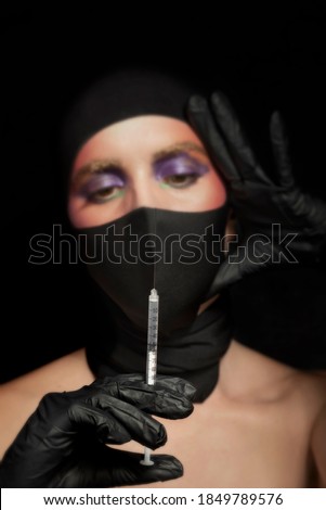 Blurred portrait in the background of a woman very well make up with a black cap and mask, in the foreground a covid vaccine injector. Artistic beauty photography. Medical artistic