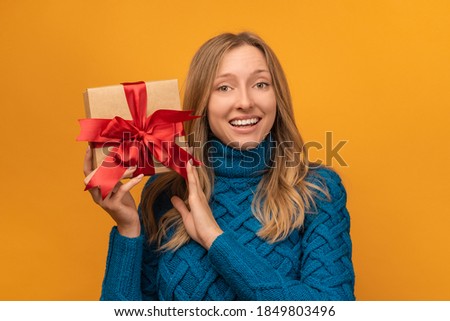 Image of charming young woman in warm knitted sweater smiling and holding gift with red ribbon. Studio shot, yellow background. New Year, Women's Day, Birthday, Holiday concept