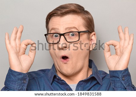 Studio close-up portrait of amazed impressed blond mature man with glasses, showing okay gestures with both hands, expressing his approval and acceptance. Headshot over gray background