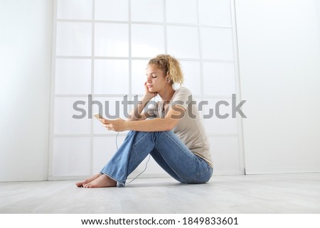 young woman with mobile phone sitting on the floor dressed casual with curly and long red hair isolated on white window background