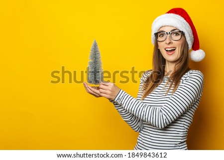 Young woman with a surprised face in a Christmas hat holds a beautiful Christmas tree in her hands on a yellow background. Banner. Christmas and New Year concept