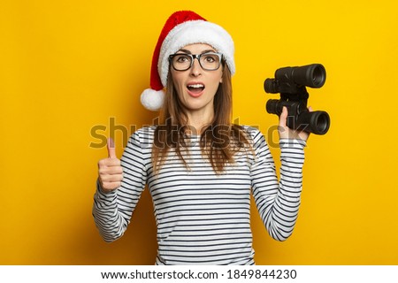 Young woman in a Santa Claus hat holds binoculars and makes a gesture All is well on a yellow background. Banner