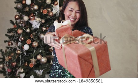 A woman shows a beautiful Christmas present against the background of a Christmas tree decorated for the holiday. Celebrating winter holidays. Receiving a Christmas surprise.
