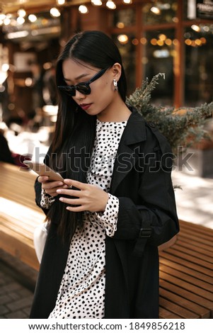 Tanned Asian woman holds phone. Brunette young lady in sunglasses, black trench coat and dress messaging outside.