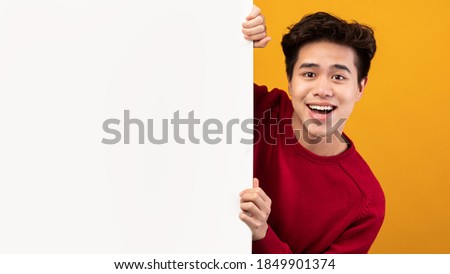 Advertising Billboard. Portrait of smiling asian guy hiding behind blank white poster with mockup template and copy space for logo, design or message. Male teen peeking out, orange studio wall Royalty-Free Stock Photo #1849901374