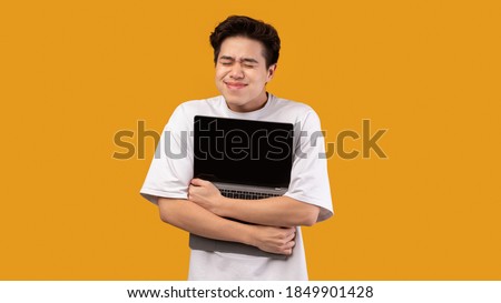 Technology Lover Concept. Happy excited asian man hugging laptop with black blank empty screen, holding it tight near chest. Geek posing isolated over orange studio background wall, copy space Royalty-Free Stock Photo #1849901428