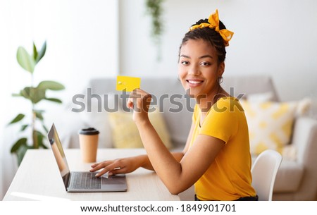 Online Shopping Concept. Side view of smiling african american woman holding credit card in hand and using laptop, ordering gifts, typing on keyboard, looking at camera, blurred background