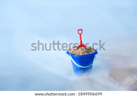 A toy beach bucket surrounded by foamy seawater at the beach, shot with slow shutter to get motion blur of the water.