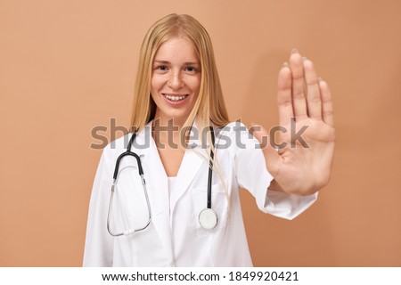 Medicine and health care concept. Portrait of adorable positive young European female doctor in white gown reaching out her hand, making stop gesture as sign of preventive measures, smiling broadly
