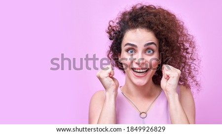 Portrait of armenian positive curly haired girl in pink dress, expressive face with bulging eyes and smile, clenched fists celebrating victory. Studio shot isolated on wall background