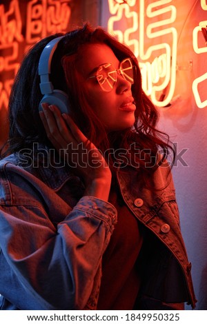 African American lady wearing headphones listening music in neon light. Cool funky young mixed race hipster listening trendy dance music stands near neon night club sign. Translation: Chicken laugh