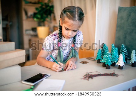 Cute girl preparing DIY gifts and signing tags to parents and family for Christmas, a green cone as if it is small toy pine tree, tags and festive boxes, handmade presents, zero waste holidays