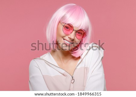 Close-up portrait of young smiling woman wearing sweatshirt, wig and eyeglasses, enjoying party and her fancy look, isolated on pink background