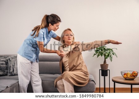 Nurse checking on blond senior woman's painful arm. Home interior.