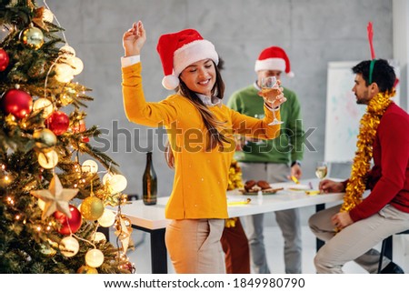 Cheerful young businesswoman with santa's hat holding champagne dancing next to christmas tree in her firm. In background are her colleagues celebrating christmas, too.