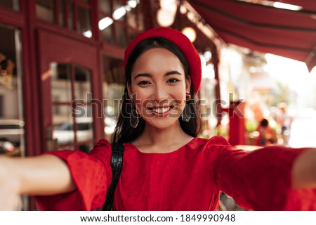 Brown-eyed brunette Asian woman in red dress, bright beret and with stylish earrings smiles widely and takes selfie in street cafe.