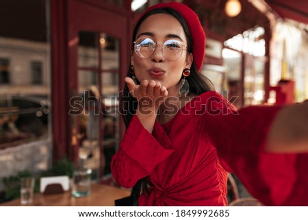 Attractive tanned woman in bright dress, red beret and eyeglasses takes selfie in street cafe and blows kiss. Elegant lady poses outside.