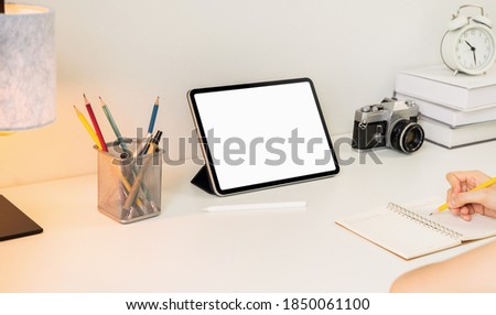 Woman hand writing on a notebook and blank white screen tablet and camera on the table.