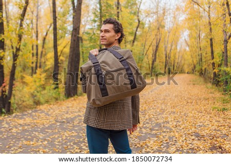 Tall handsome man dressed in a brown jacket walking with a bag on the autumn alley