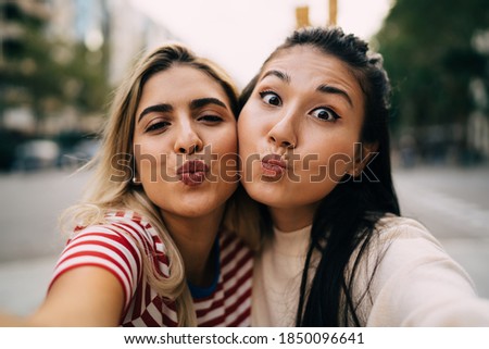 Close up portrait of Hispanic and Asian female friends making duck face and looking at camera spending togetherness leisure time, multicultural vloggers showing kiss during selfie photographing
