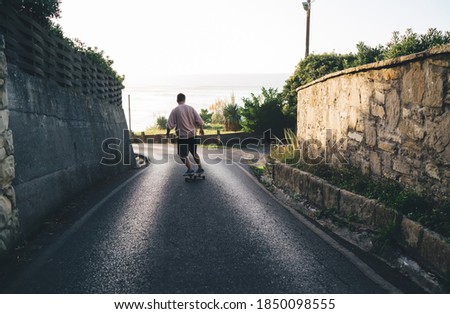 Back view of unrecognizable man skate rider in casual clothes training alone on empty asphalt roadway on sunny day among green plants