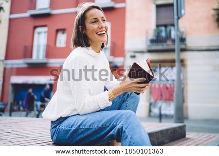 Low angle of smiling stylish woman in jeans with interesting book having fun on staircase of city park during weekend