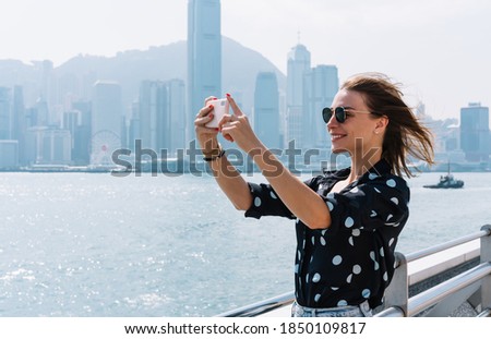 Happy blonde female blogger shooting video on mobile phone spending time in journey on sunny summer day, positive woman 20s influencer making photo via app on smartphone on bridge in downtown