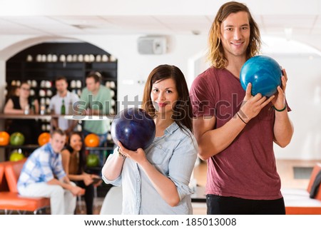 Portrait of confident man and woman holding bowling balls with friends in background