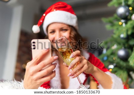 The celebration of new year and Christmas on the distance of the concept. A young woman in a Santa suit and hat is holding a smartphone against the background of a decorated fir tree.