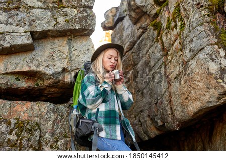 charming blonde female is drinking hot tea during hiking, stand near big rocks in mountains, travel and adventure concept