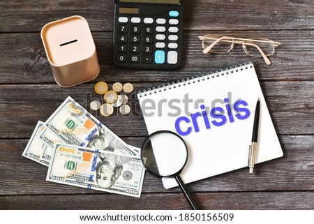CRISIS. The word CRISIS is written in a white notebook on a wooden background near a black magnifier, glasses, money, calculator. Business concept. Flat lay.